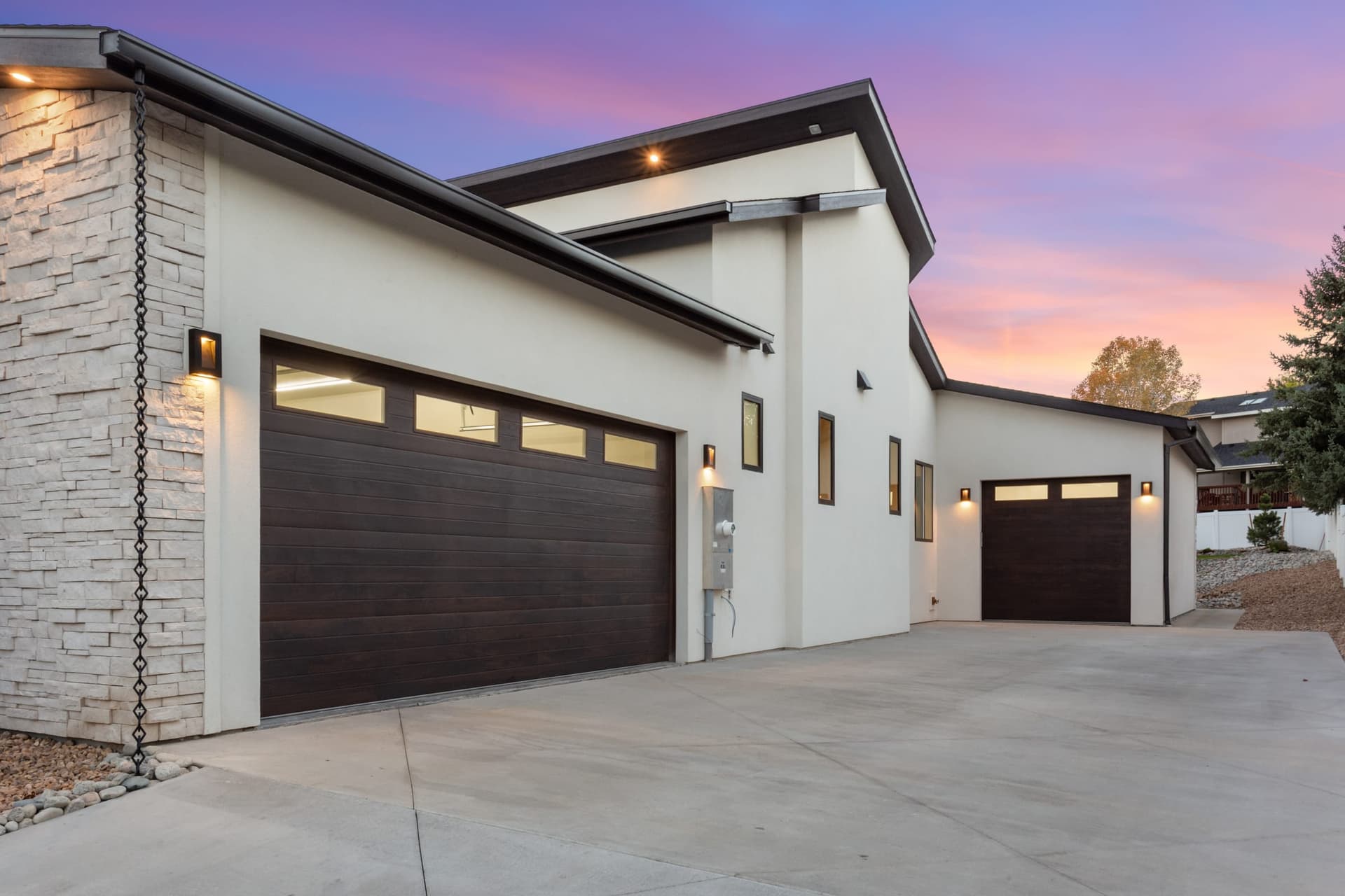 Garage elevation at twilight with modern wall sconces, cantilevered overhangs, and dark garage doors