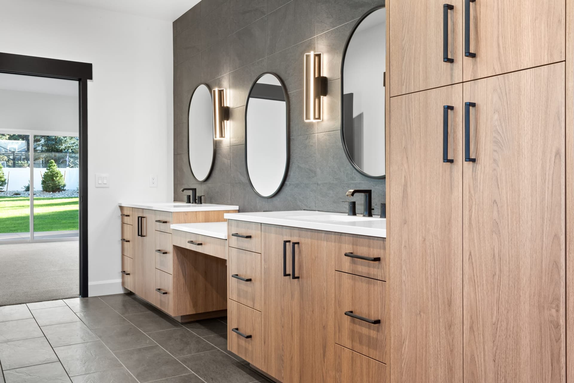 Primary bath vanity detail — warm wood cabinetry, matte black hardware, oval mirrors, brass cylindrical wall sconces